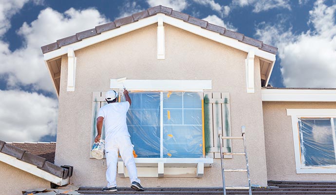A professional painter in white clothes uses a roller to paint the trim of a house window from a ladder, with the windows covered in plastic