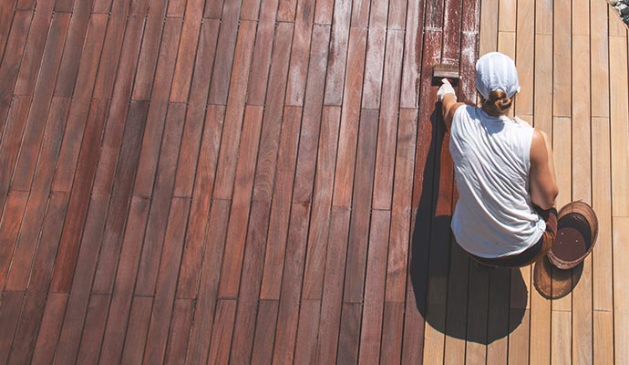 An overhead view of a person on a wooden deck, using a brush to apply a dark brown stain to the light-colored wooden planks.