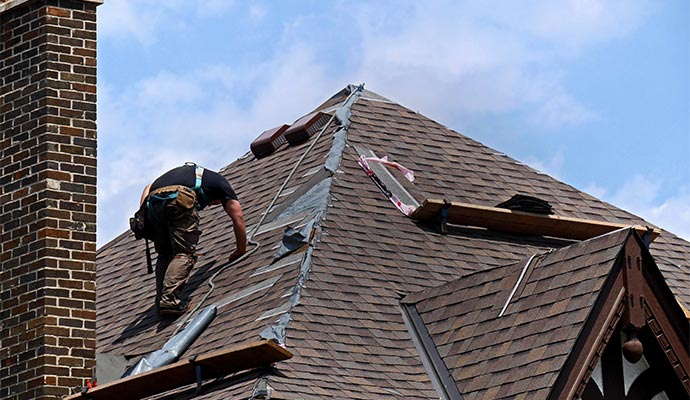 A roofer wearing a safety harness and tool belt works on a steep residential roof with brown shingles