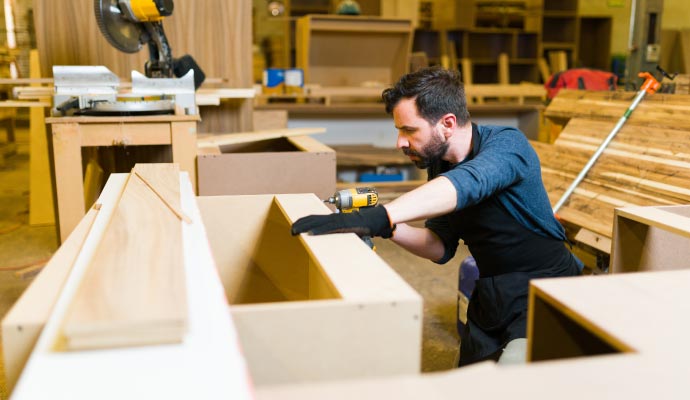 A carpenter in a workshop uses a power drill to assemble custom cabinets from wood and MDF.