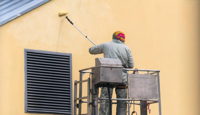 Commercial building exterior painting A painter on a vertical platform lift uses a roller on an extension pole to paint the yellow exterior wall of a commercial building.