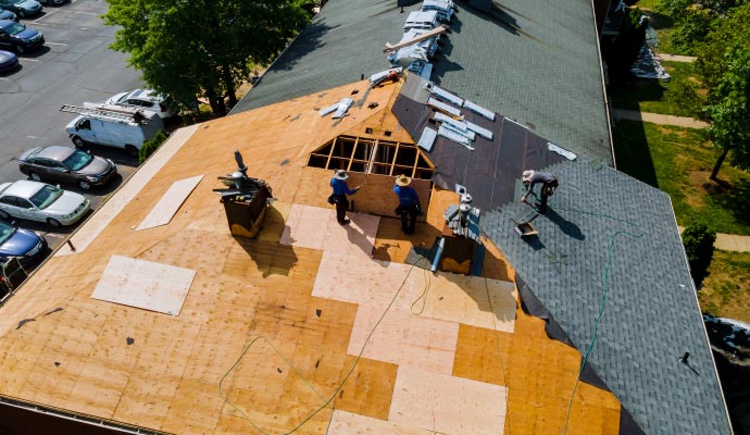 An aerial view of a large roof replacement project, showing workers tearing off old shingles and installing new plywood sheathing and underlayment.