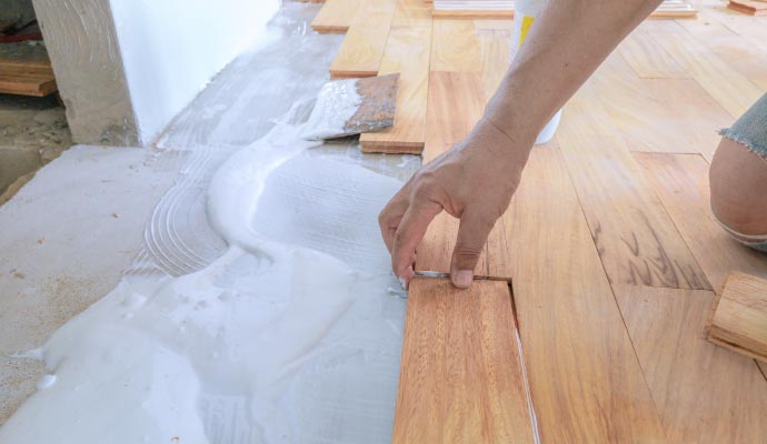A close-up of a worker's hand laying a hardwood floor plank into a thick layer of white adhesive on a concrete subfloor.