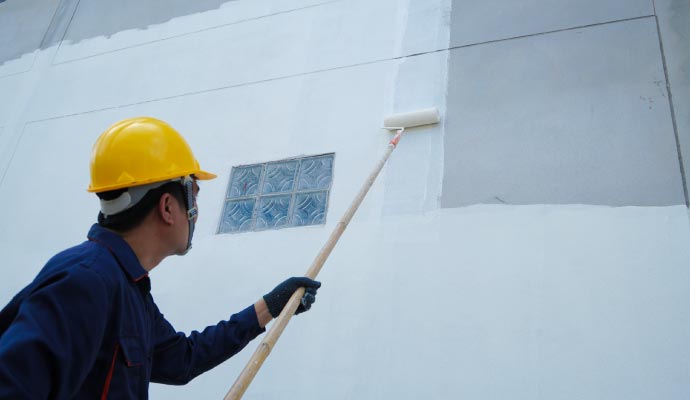 Commercial exterior painting: primer coat A painter wearing a yellow hard hat uses a roller on an extension pole to apply white primer to the large grey exterior wall of a commercial building.