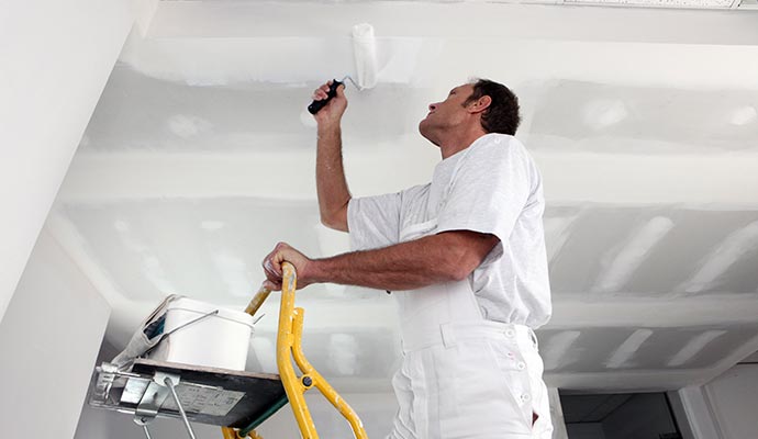 A painter painting a drywall ceiling using a roller in an interior room