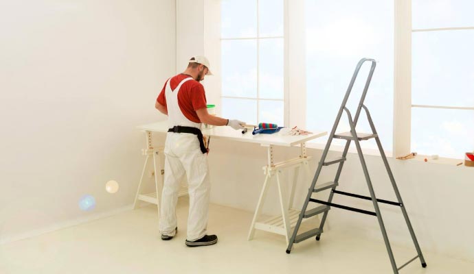 A painter in white overalls stands at a temporary work table, preparing his tools in a bright, white room with a large window.