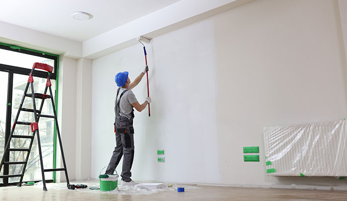 A painter in a hard hat painting a white wall with a roller.
