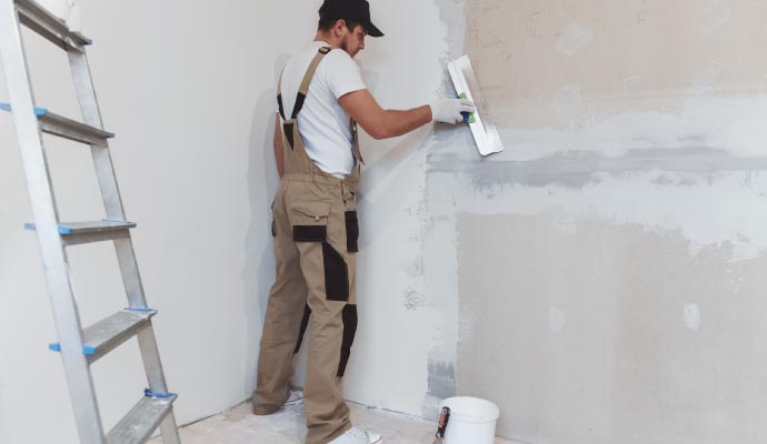 A worker in overalls uses a long trowel to apply a thin skim coat of plaster to a wall during a renovation.