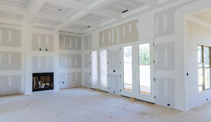 A large room under construction showing the drywall finishing stage, with a coffered ceiling, a fireplace, and a wall of French doors.