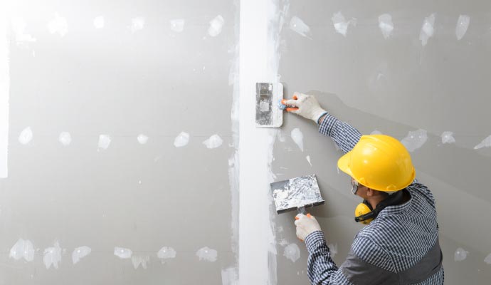 A worker in a yellow hard hat uses taping knives to apply joint compound to an inside corner of a drywall installation.