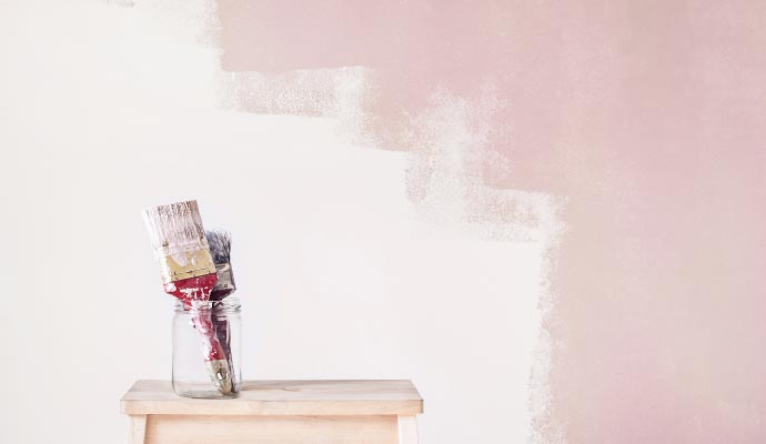 Used paintbrushes resting in a glass jar on a wooden stool, in front of a wall partially painted a dusty pink color.