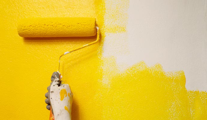 A close-up of a gloved hand using a paint roller to apply bright yellow paint over a white wall.