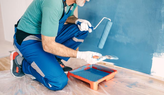 A painter in blue overalls crouches down to paint a wall blue, using a small paint roller and a paint tray.